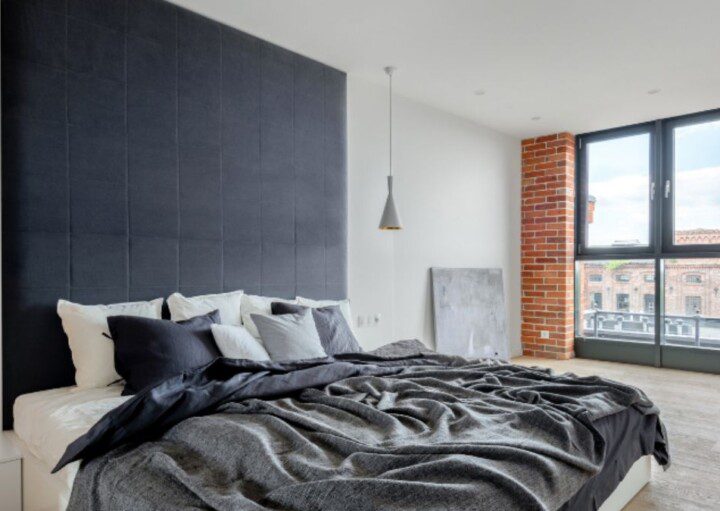 A imagem mostra uma cama king size em quarto amplo em decoração industrial, com lâmpadas dos dois lados e janelas abertas de vidro.