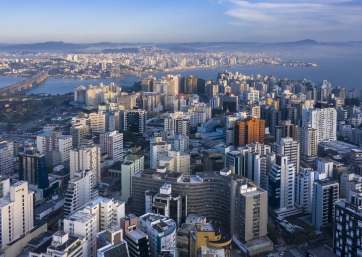 Imagem de edifícios do centro de Florianópolis vistos do alto com ponte suspensa a cabo ao fundo para ilustrar matéria sobre as melhores cidades de Santa Catarina