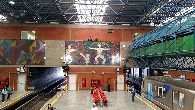 Uma plataforma de estação de metrô com assentos vermelhos, pessoas esperando, murais abstratos em paredes de tijolos e uma passarela de pedestres de metal azul acima. Um trem prateado do metrô está parado no lado direito da imagem.