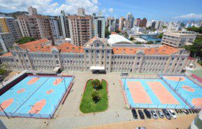 Foto que ilustra matéria sobre escolas particulares em Florianópolis mostra a fachada do Colégio Catarinense. À frente do prédio, há quadras poliesportivas. E atrás dele há prédios residenciais da cidade. Mais ao fundo, o céu azul de um dia ensolarado.