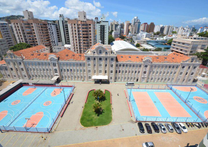 Foto que ilustra matéria sobre escolas particulares em Florianópolis mostra a fachada do Colégio Catarinense. À frente do prédio, há quadras poliesportivas. E atrás dele há prédios residenciais da cidade. Mais ao fundo, o céu azul de um dia ensolarado.