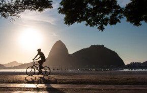 Foto que ilustra matéria sobre morar no Rio de Janeiro mostra a silhueta do morro do Pão de Açúcar. Ao lado esquerdo dele, aparece o sol baixo. E em primeiro plano, aparece a silhueta de um ciclista.