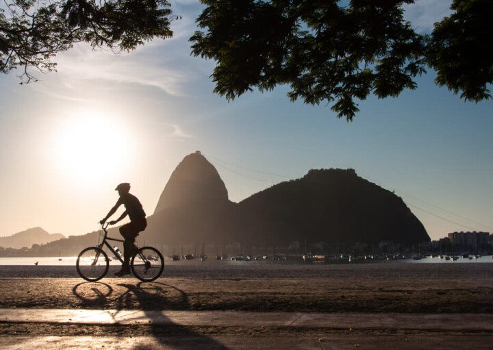 Foto que ilustra matéria sobre morar no Rio de Janeiro mostra a silhueta do morro do Pão de Açúcar. Ao lado esquerdo dele, aparece o sol baixo. E em primeiro plano, aparece a silhueta de um ciclista.