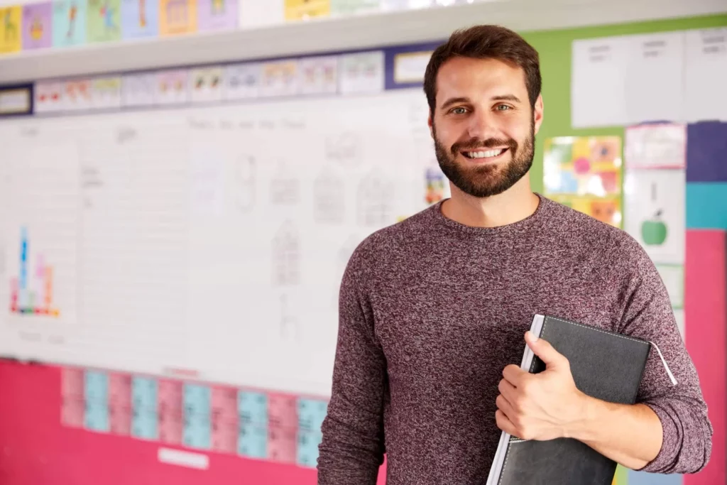 Imagem de um homem sorridente com barba segura uma pasta preta, em pé em uma sala de aula colorida, com um quadro branco e trabalhos artísticos de alunos ao fundo