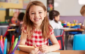 Imagem de uma menina sorridente, vestindo uma camisa xadrez vermelha e branca, senta-se em uma mesa de sala de aula segurando um lápis sobre um desenho. Outras crianças trabalham ao fundo com materiais escolares coloridos sobre a mesa