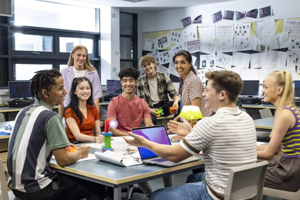 Imagem de um grupo diversificado de alunos senta-se ao redor da mesa de uma sala de aula, sorrindo e conversando. Papéis, laptops e materiais de projeto estão espalhados sobre a mesa, com pôsteres e diagramas nas paredes atrás deles