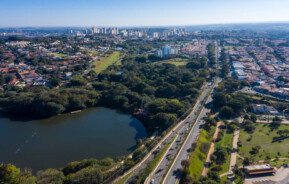 Foto que ilustra matéria sobre morar em Campinas mostra uma visão do alto de uma parte da cidade, com destaque para a Lagoa do Taquaral, que aparece no canto inferior esquerdo. No meio da tela, algumas árvores, mais atrás muitas casas e no horizonte mais ao fundo os prédios mais altos, com um céu azul de fundo.