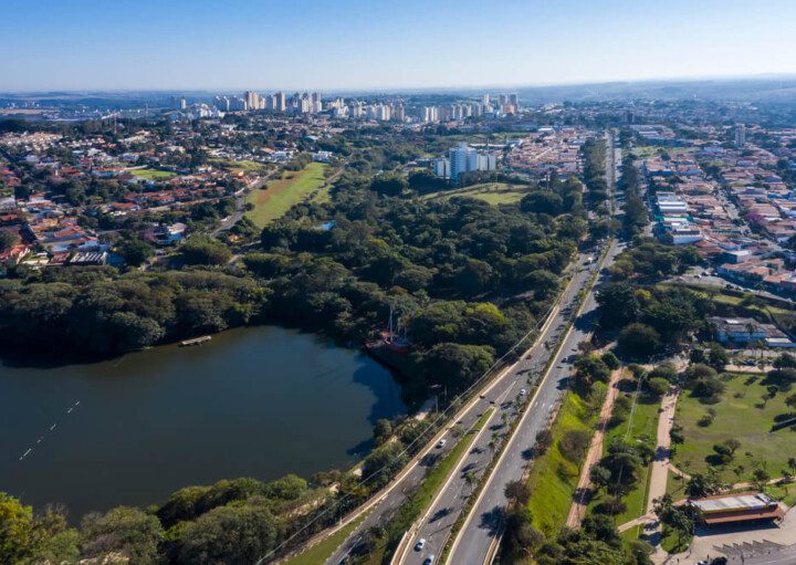 Foto que ilustra matéria sobre morar em Campinas mostra uma visão do alto de uma parte da cidade, com destaque para a Lagoa do Taquaral, que aparece no canto inferior esquerdo. No meio da tela, algumas árvores, mais atrás muitas casas e no horizonte mais ao fundo os prédios mais altos, com um céu azul de fundo.