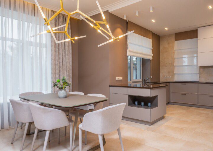 Cozinha moderna e sala de jantar minimalista com tons de bege, uma mesa retangular com seis cadeiras claras, grandes janelas com cortinas transparentes, um lustre geométrico dourado exclusivo e armários minimalistas com um vaso de flores sobre a mesa.