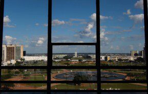 Foto que ilustra matéria sobre bairros de Brasília mostra a Esplanada dos Ministérios vista de dentro de um prédio localizado de frente para a região. Em primeiro plano, aparecem, em sombras, a estrutura das janelas do prédio de onde a foto foi tirada. Bem ao fundo, aparece o Congresso Nacional.