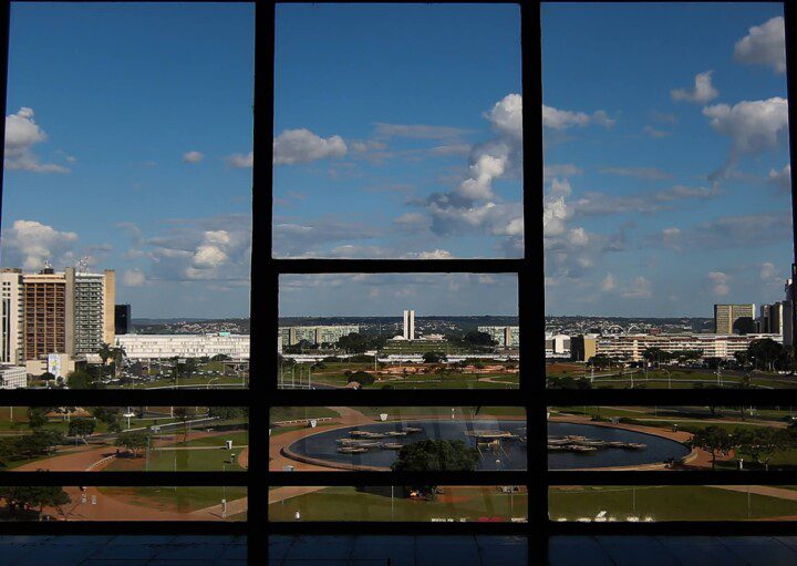 Foto que ilustra matéria sobre bairros de Brasília mostra a Esplanada dos Ministérios vista de dentro de um prédio localizado de frente para a região. Em primeiro plano, aparecem, em sombras, a estrutura das janelas do prédio de onde a foto foi tirada. Bem ao fundo, aparece o Congresso Nacional.