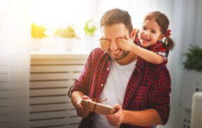 Foto que ilustra matéria sobre decoração para Dia dos Pais mostra um homem sorridente segurando um embrulho pequeno de presente nas mãos enquanto uma menina, também sorridente, está atrás e tampando os olhos dele com as mãos