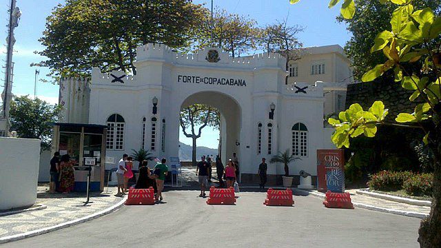 Pessoas param perto da entrada do Forte de Copacabana, um forte branco histórico e um dos museus mais notáveis do Rio de Janeiro, com árvores e um céu limpo ao fundo. Barricadas laranjas bloqueiam o acesso de veículos.