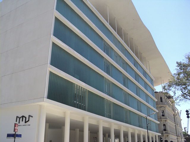 Um edifício moderno com altas colunas brancas, grandes janelas verticais de vidro e um telhado plano e inclinado. Reminiscente dos museus do Rio de Janeiro, seu design minimalista convive com uma estrutura mais tradicional.