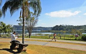 Foto que ilustra matéria sobre o que fazer em Jundiaí mostra uma paisagem do Parque da Cidade, com uma mulher sentada em um banco enquanto olha uma câmera fotográfica à esquerda, em primeiro plano, uma pista de corrida, com um homem se exercitando, e ao fundo um espelho d’água seguido de árvores em um dia de céu azul e poucas nuvens.