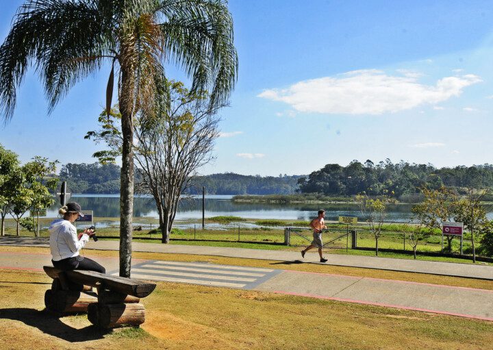 Foto que ilustra matéria sobre o que fazer em Jundiaí mostra uma paisagem do Parque da Cidade, com uma mulher sentada em um banco enquanto olha uma câmera fotográfica à esquerda, em primeiro plano, uma pista de corrida, com um homem se exercitando, e ao fundo um espelho d’água seguido de árvores em um dia de céu azul e poucas nuvens.