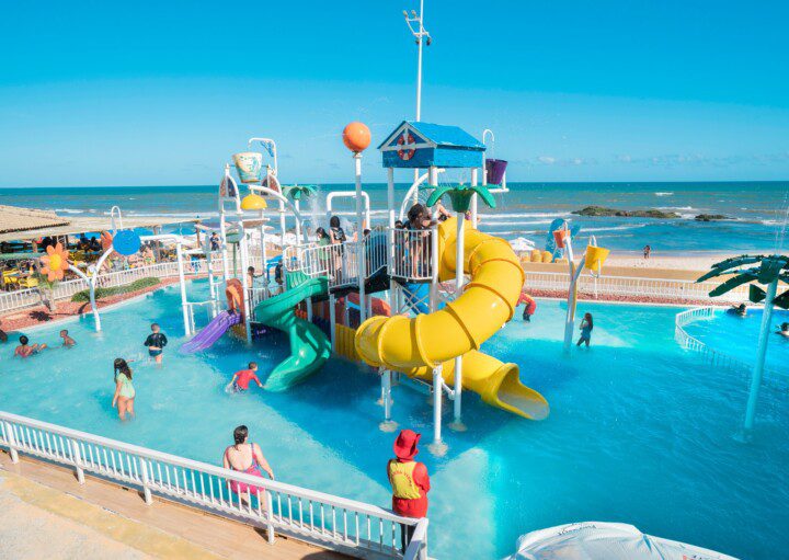 Foto que ilustra matéria sobre parque aquático em Salvador mostra o brinquedo aquático do Salvador Beach Club, uma estrutura com escorregadores coloridos em meio a uma piscina, com uma praia ao fundo.