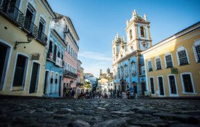 Foto que ilustra matéria sobre o Pelourinho, em Salvador, mostra uma das ruas de pedra da região em um dia de céu azul. A foto foi tirada de um ângulo de baixo para cima, de forma que o chão aparece bem. À esquerda e à direita aparecem os sobrados coloridos da região, com destaque, à direita, para a Igreja do Rosário dos Pretos.