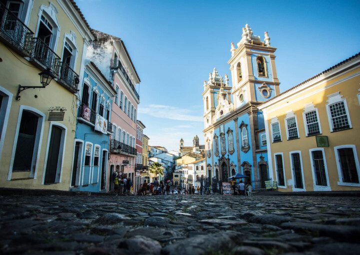 Foto que ilustra matéria sobre o Pelourinho, em Salvador, mostra uma das ruas de pedra da região em um dia de céu azul. A foto foi tirada de um ângulo de baixo para cima, de forma que o chão aparece bem. À esquerda e à direita aparecem os sobrados coloridos da região, com destaque, à direita, para a Igreja do Rosário dos Pretos.