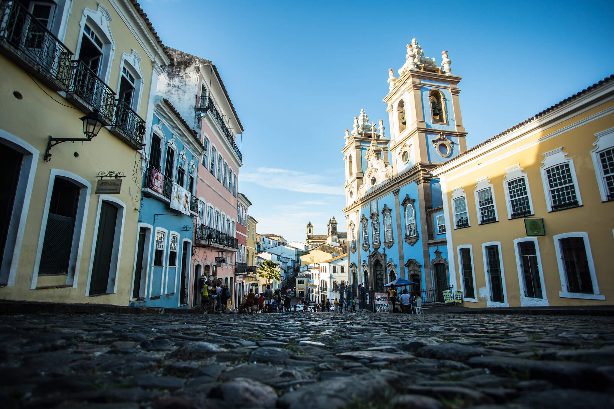 Foto que ilustra matéria sobre o Pelourinho, em Salvador, mostra uma das ruas de pedra da região em um dia de céu azul. A foto foi tirada de um ângulo de baixo para cima, de forma que o chão aparece bem. À esquerda e à direita aparecem os sobrados coloridos da região, com destaque, à direita, para a Igreja do Rosário dos Pretos.