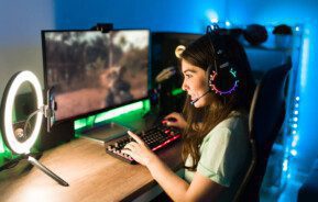 Foto que ilustra matéria sobre sala gamer mostra uma jovem sorridente, sentada de lado, no canto direito da tela, e diante de um monitor com um jogo rodando. Ela está com a mão esquerda em um teclado, a mão direita em um mouse e tem um fone nos ouvidos.