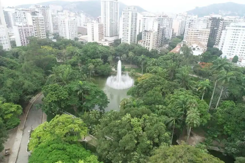 Imagem da vista aérea do Campo de São Bento, um Parque em Niterói, mostra densas árvores verdes e um lago central com uma fonte de água alta cercado por prédios altos 