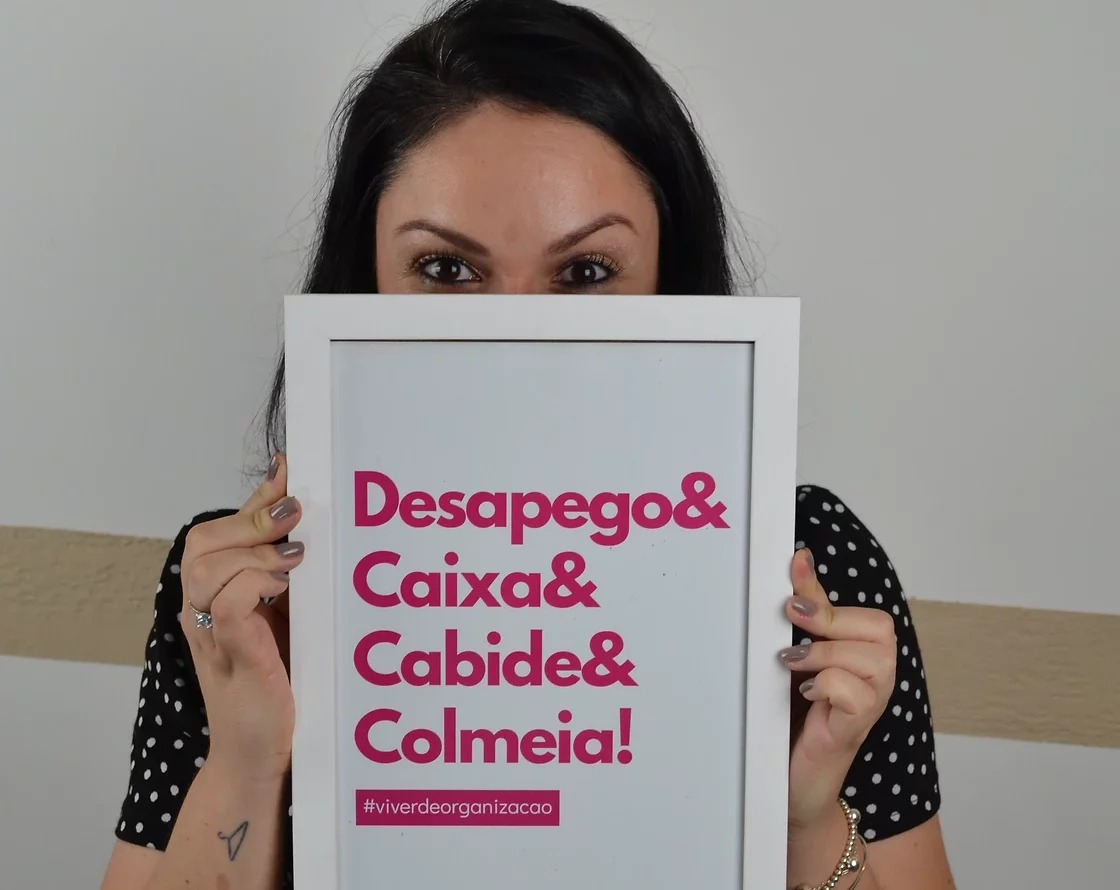 Foto que ilustra bloco sobre a autora mostra a Personal Organizer Aline Delavy segurando um quadro que diz "Desapego e Caixa e Cabide e Colmeia"em frente ao rosto.