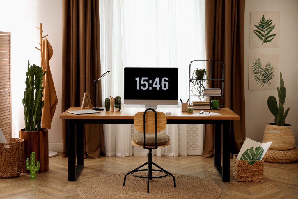 Na foto há um escritório com: mesa de trabalho, cadeira, computador, cabide, cacto, cestos organizadores e itens de escritório como canetas e blocos de notas.