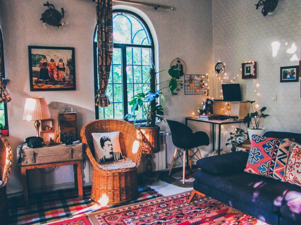 A imagem mostra uma sala com decoração maximalista. Nela há poltrona, sofá, mesa de estudos e cadeira. Tem também muitos elementos decorativos como almofadas, tapetes, quadros, abajur, entre outros.