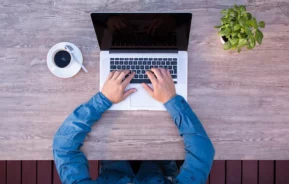 Imagem que ilustra matéria sobre comprar ou investir em fundos imobiliários mostra um homem sentado em frente a um computador com um café do lado esquerdo e um vaso de planta do lado direito.