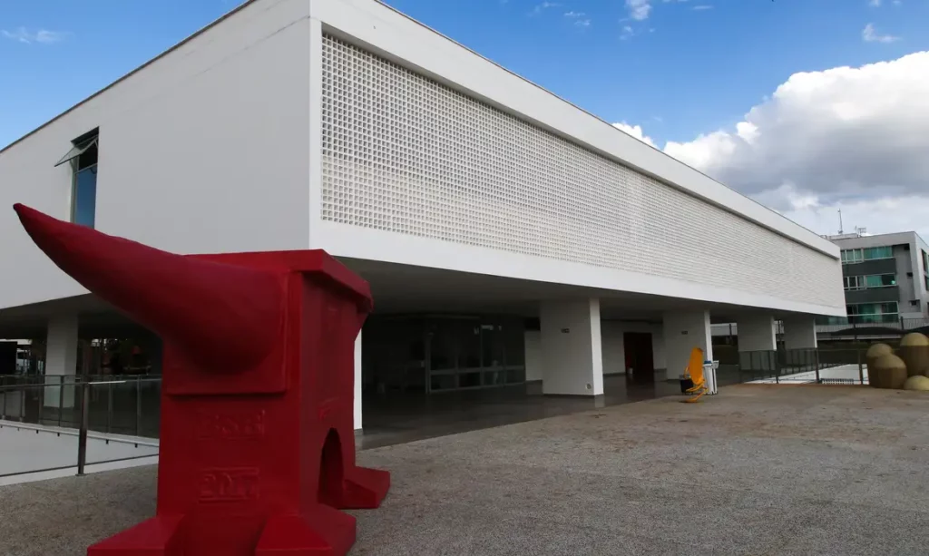 Imagem do Museu de Arte de Brasília (MAB), um prédio branco e moderno com uma fachada em forma de grade; em primeiro plano, uma escultura abstrata vermelha brilhante com uma saliência semelhante a um chifre. O céu está parcialmente nublado