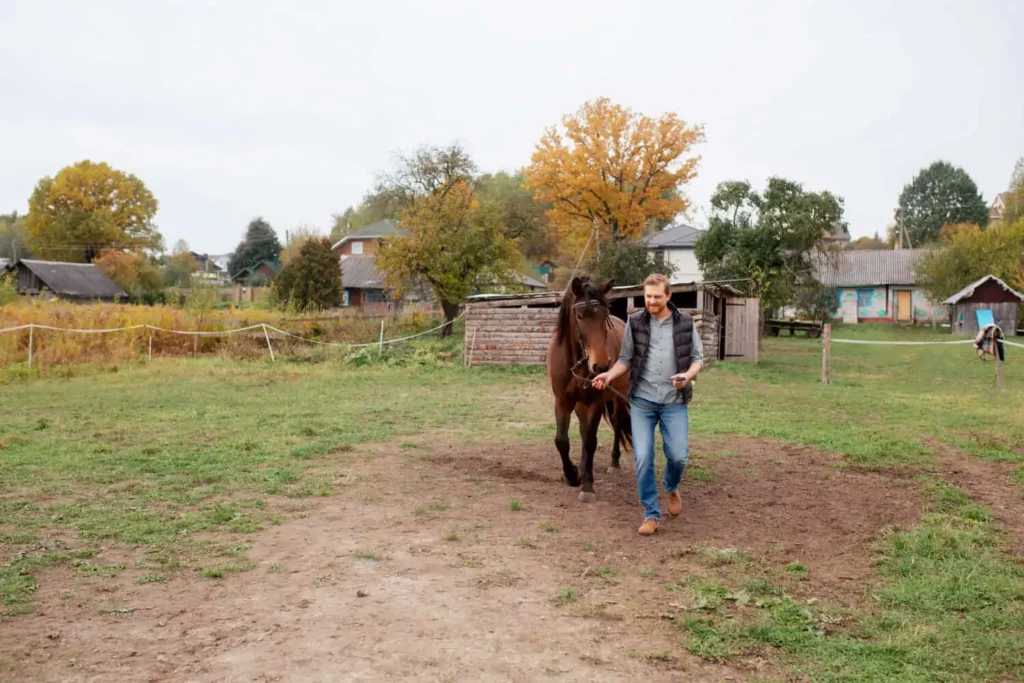 Imagem de um homem vestindo jeans e jaqueta cinza caminha ao lado de um cavalo marrom em uma área rural gramada, com galpões e árvores ao fundo em um dia nublado, para ilustrar matéria sobre o que significa NIRF