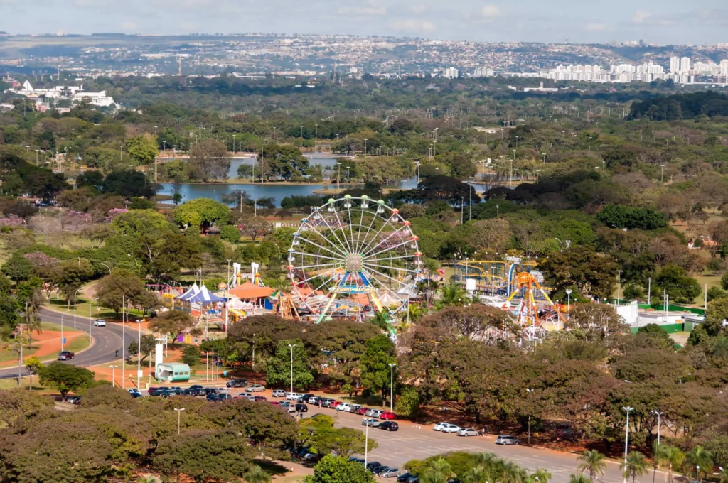 Imagem da vista aérea do Parque da Cidade de Brasília mostra um parque de diversões colorido com uma roda-gigante cercada por árvores perto de um lago