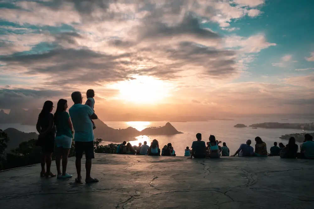 Imagem de pessoas em pé e outras sentadas no mirante do Parque da Cidade, em Niterói, observando o pôr do sol sobre montanhas, ilhas e água sob um céu parcialmente nublado.