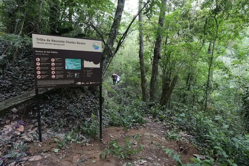 Imagem de uma trilha florestal no Parque Estadual Serra da Tiririca, em Niterói, mostra uma placa onde se lê Trilha da Nascente Charles Darwin. Árvores densas e verdes cercam o caminho e uma pessoa é vista caminhando à distância ao longo da trilha cênica.