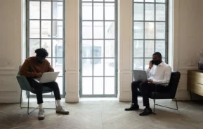 Foto que ilustra matéria mostra uma sala com dois homens de máscara sentados em cadeiras distintas, ambos estão mexendo em seus respectivos computadores.