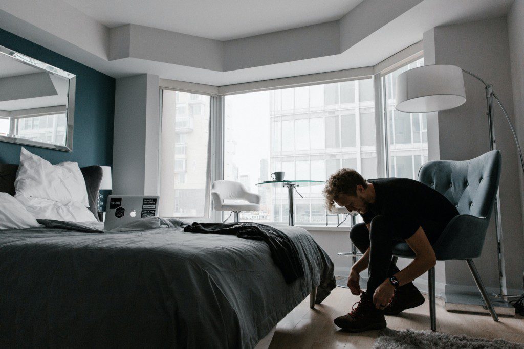 Foto de um quarto moderno em tons escuros. Na imagem há uma cama de casal, uma poltrona com design inovador e uma luminária de chão em metal. Há também um homem calçando o tênis na poltrona.