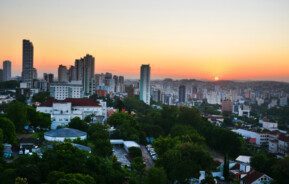 Foto que ilustra matéria sobre bairros de Novo Hamburgo mostra uma vista aérea da cidade com o sol se pondo no horizonte ao fundo. A imagem mostra, em primeiro plano, algumas árvores e casas e mais ao centro e à esquerda alguns prédios.