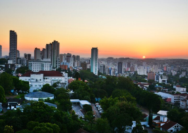 Foto que ilustra matéria sobre bairros de Novo Hamburgo mostra uma vista aérea da cidade com o sol se pondo no horizonte ao fundo. A imagem mostra, em primeiro plano, algumas árvores e casas e mais ao centro e à esquerda alguns prédios.