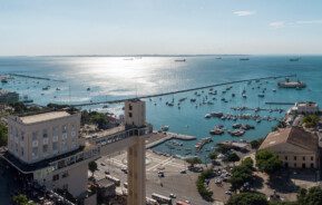 Foto que ilustra matéria sobre custo de vida em Salvador mostra uma visão do alto do Elevador Lacerda, que aparece no canto inferior esquerdo da imagem, que é boa parte ocupada pelo espelho d’água da Baía de Todos os Santos, onde aparece vários barcos. Ao fundo, um céu azul com o dia claro.