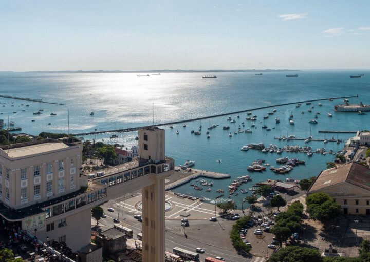 Foto que ilustra matéria sobre custo de vida em Salvador mostra uma visão do alto do Elevador Lacerda, que aparece no canto inferior esquerdo da imagem, que é boa parte ocupada pelo espelho d’água da Baía de Todos os Santos, onde aparece vários barcos. Ao fundo, um céu azul com o dia claro.