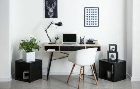Foto que ilustra matéria sobre decoração de escritório moderno mostra uma mesa de computador encostada em uma parede branca com dois pequenos quadros acima. Em cima da mesa há um notebook, uma luminária e materiais de escritório, como canetas e papéis. Dos dois lados da mesa há dois nichos pretos em forma de cubo no chão. O da esquerda tem um vaso com uma planta.