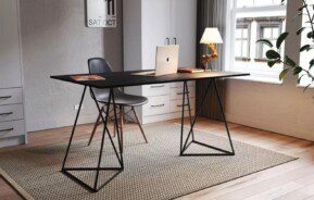 Um home office moderno com decoração de escritório moderna apresenta uma mesa preta, cadeira cinza, laptop, luminária, livros e tapete. Uma grande janela preenche o espaço com luz natural; armários brancos e uma planta verde iluminam o fundo.