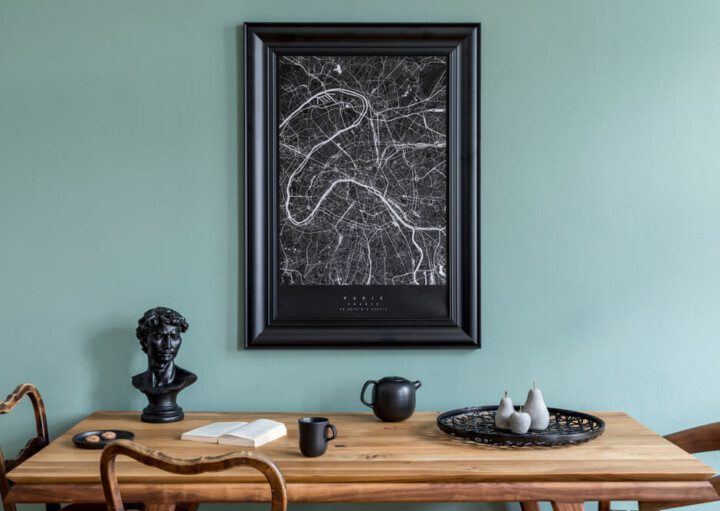 Foto que ilustra matéria sobre decoração de mesa mostra uma mesa de jantar de madeira clara encostada em uma parede verde. Em cima da mesa há objetos decorativos como uma pequena estátua, além de um livro aberto, um bule e uma xícara. Na parede há um quadro.