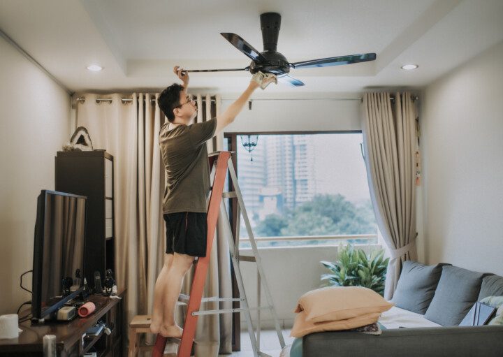 Imagem de um homem em cima de uma escada limpando um ventilador de teto com um pano.