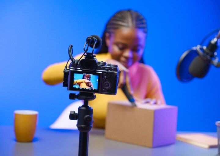 Foto que ilustra matéria sobre como montar um estúdio em casa mostra uma mulher abrindo uma caixa com um fundo azul enquanto filma o processo e fala em um microfone