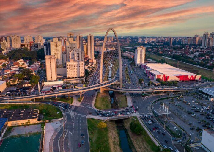 Foto que ilustra matéria sobre morar em São José dos Campos mostra uma vista aérea de uma ponte estaiada localizada na cidade, com diversas avenidas que se cruzam do meio para baixo da imagem. Nos cantos superiores, há vários prédios E mais ao fundo, um céu ao entardecer com nuvens avermelhadas.