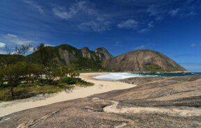 Foto que ilustra matéria sobre o que fazer em Niterói mostra uma vista panorâmica de um trecho da praia de Itacoatiara com uma pequena facha de areia, um pedaço das ondas do mar aparecendo e um grande maciço de pedra ao fundo em um dia de céu azul