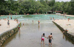 Foto que ilustra matéria sobre parques em Brasília mostra a entrada de uma das piscinas do Parque Nacional de Brasília, repleta de pessoas se banhando e com várias árvores ao fundo.