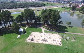 Foto que ilustra matéria sobre parques em São José dos Pinhais mostra a visão aérea do parque municipal da cidade, com um espaço de areia dentro de um gramado, onde se encontram aparelhos de ginástica, árvores e um lago mais ao fundo.